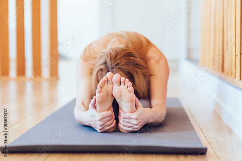 Close up of young woman practicing yoga, sitting in Head to Knee Forward Bend exercise, Janu Sirsasana pose, working out, wearing sportswear, home interior background photo