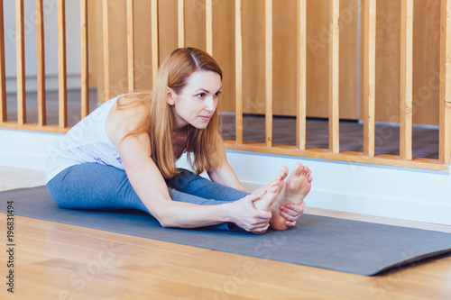 Middle aged blond woman practicing yoga, sitting in Seated forward bend exercise, paschimottanasana pose, working out, wearing sportswear, grey pants, indoor, home interior wooden background,side view photo