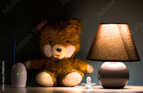 Bedside lamp, teddy bear, pacifier, baby sound monitor in baby room at night.