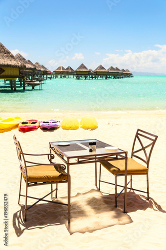 Dining table with two chairs on sandy beach by tropical sea side, with colourful kayaks and water villas background