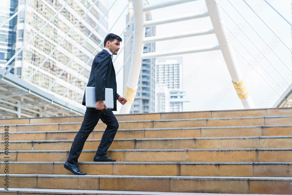 Business man celebrating success. Young businessman walking at stairs and hold laptop computer while standing outdoors with office building in the background. Business man celebrating success. Young businessman walking at stairs and hold laptop computer while standing outdoors with office building in the background.