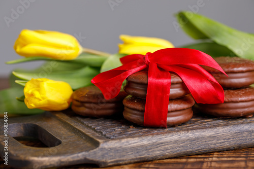 Yellow tulips and chocolate cookies tied with satin red ribbon on a wooden background. Flowers and treats on a beautiful wooden board with a pattern.