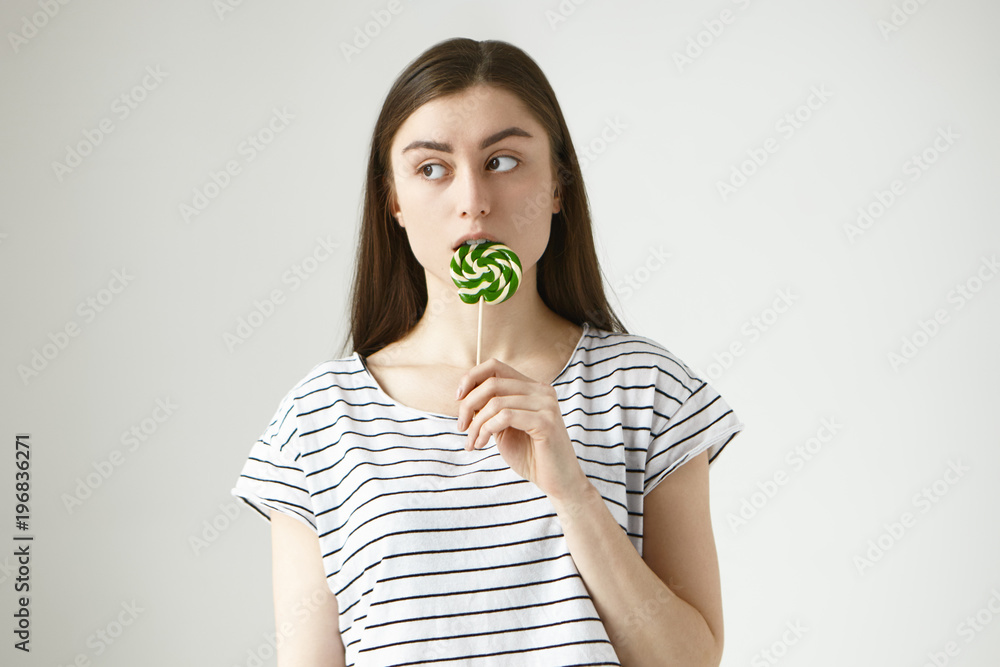Horizontal picture of brunette young female on strict diet licking colorful yellow green lollipop in secrecy, looking sideways, feeling worried of being caught eating unhealthy sugar hard candy Horizontal picture of brunette young female on strict diet licking colorful yellow green lollipop in secrecy, looking sideways, feeling worried of being caught eating unhealthy sugar hard candy