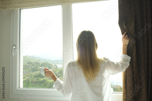 New day new life concept. Young fit woman standing by big window view holding morning coffee cup. Attractive female wearing tight shirt, sexy translucent nightie bask in sun. Hands close up background © Evrymmnt