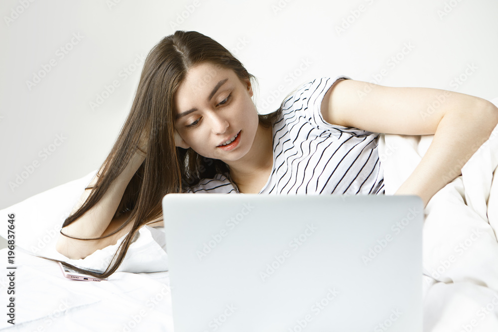 Picture of beautiful student girl relaxing at home on weekend morning, lying in bed in front of open laptop, having joyful look while watching movie online. Leisure, relaxation and electronic devices Picture of beautiful student girl relaxing at home on weekend morning, lying in bed in front of open laptop, having joyful look while watching movie online. Leisure, relaxation and electronic devices