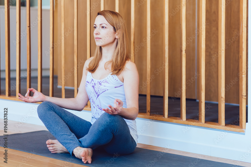 Full body portrait of a relaxed candid caucasian woman practicing yoga sitting on the floor in the empty living room at home. New day, morning routine concept. Full body portrait of a relaxed candid caucasian woman practicing yoga sitting on the floor in the empty living room at home. New day, morning routine concept.