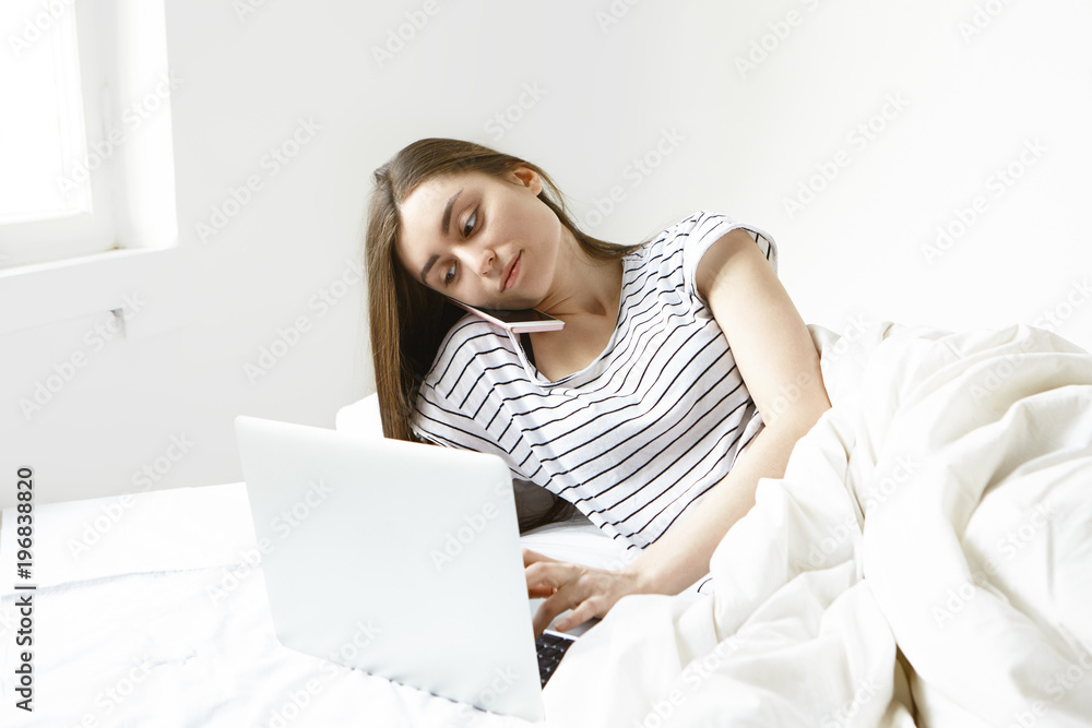 Attractive young dark haired woman in striped t-shirt multitasking while lying in white bed, keyboarding using open laptop and speaking on smart phone. People, modern gadgets and communication Attractive young dark haired woman in striped t-shirt multitasking while lying in white bed, keyboarding using open laptop and speaking on smart phone. People, modern gadgets and communication