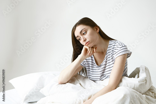 Horizontal isolated picture of upset beautiful young woman with long brown hair sitting on bed, having pensive look, unwilling to go to work, feeling sick and tired of her boring monotonous life