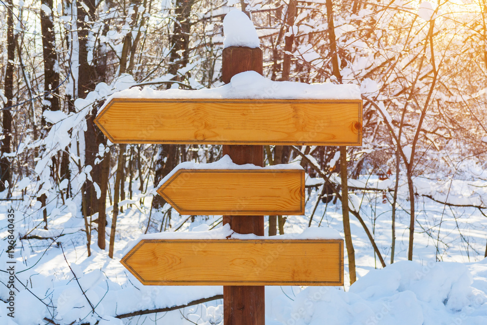 Naklejka premium Blank wooden directional arrows on a post, covered by snow in the winter forest illuminated by sunlight (toned)