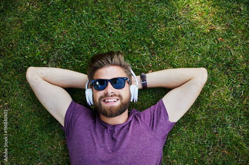 Happy man listening to music while lying on the grass outdoors and wearing sunglasses