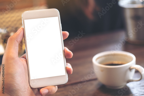 Mockup image of a hand holding white mobile phone with blank desktop screen and coffee cup on wooden table in modern cafe
