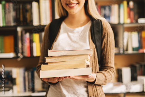 Close up view of a book stack in hands of a smiling girl with the backpack in the library.