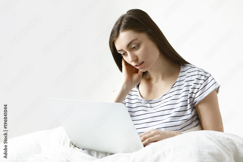 Casually dressed young European female freelancer using generic laptop for distant work, sitting on white bed and looking at screen with focused expression. Modern technologies and lifestyle Casually dressed young European female freelancer using generic laptop for distant work, sitting on white bed and looking at screen with focused expression. Modern technologies and lifestyle