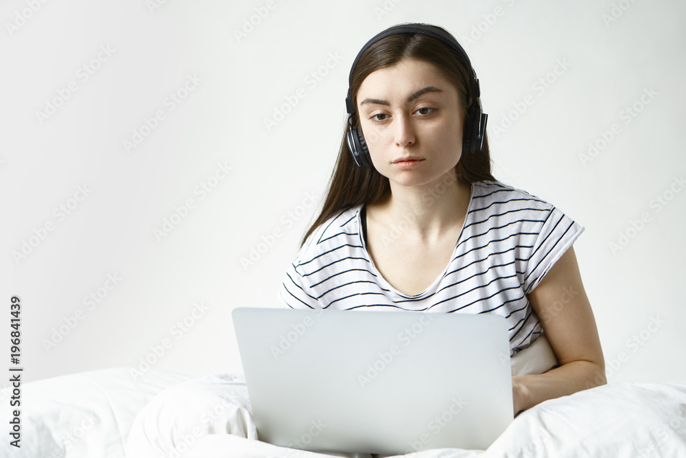 Indoor shot of beautiful brunette female student in striped t-shirt doing homework in her room, sitting on white bed with laptop and headphones, having serious concentrated facial expression Indoor shot of beautiful brunette female student in striped t-shirt doing homework in her room, sitting on white bed with laptop and headphones, having serious concentrated facial expression