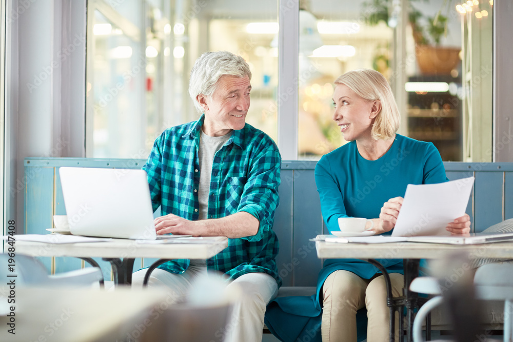 Fototapeta premium Two happy aged employees sitting by tables in cafe and looking at one another during talk