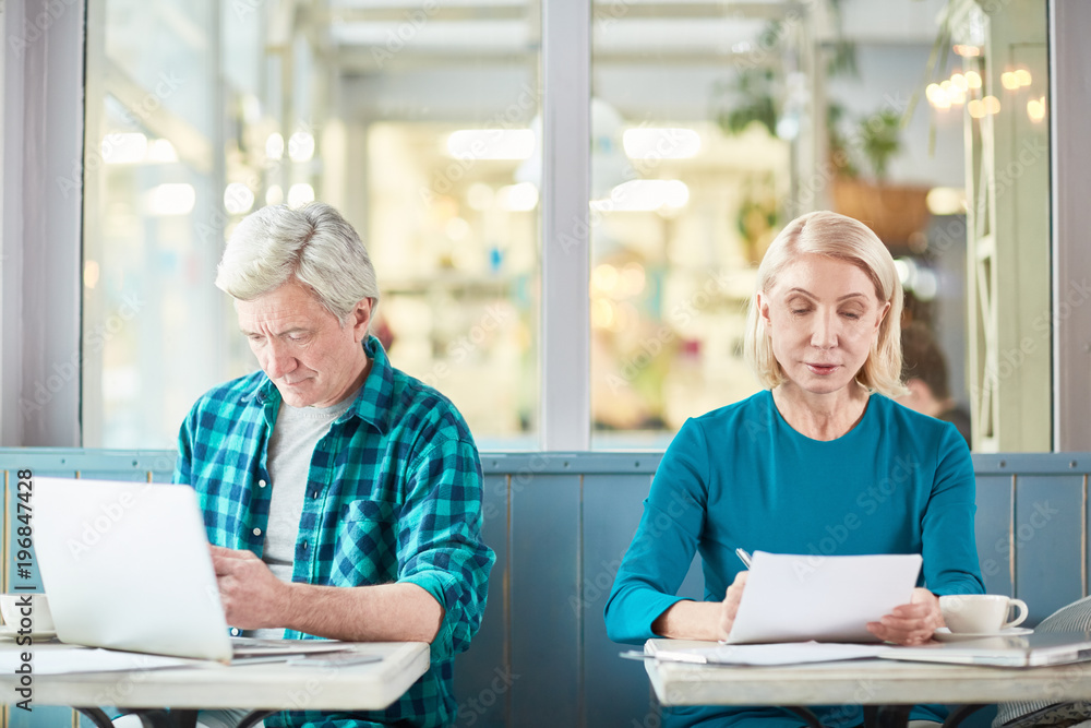 Mature man browsing in the net while analyzing data and aged woman reading financial papers while both sitting in cafe Mature man browsing in the net while analyzing data and aged woman reading financial papers while both sitting in cafe