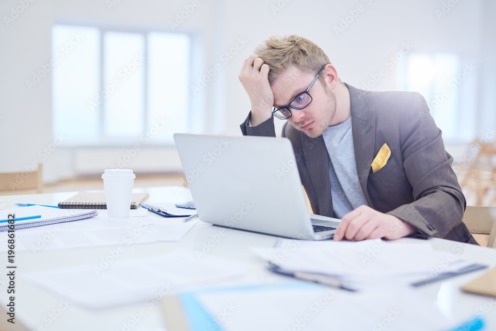 Puzzled young manager sitting by table in front of laptop and reading online data or statistics Puzzled young manager sitting by table in front of laptop and reading online data or statistics