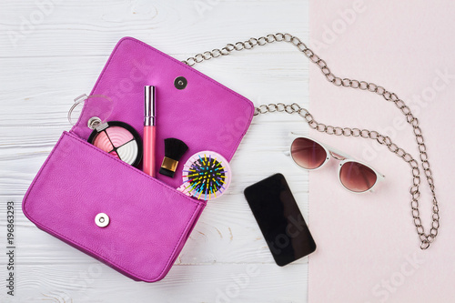 Fashion woman handbag with makeup accessories. Stylish woman essentials, cosmetics, cellphone, sunglasses on wooden background, top view.