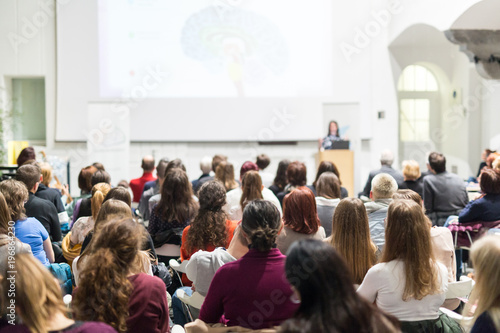 Female speaker giving presentation in lecture hall at university workshop. Audience in conference hall. Rear view of unrecognized participant in audience. Scientific conference event.