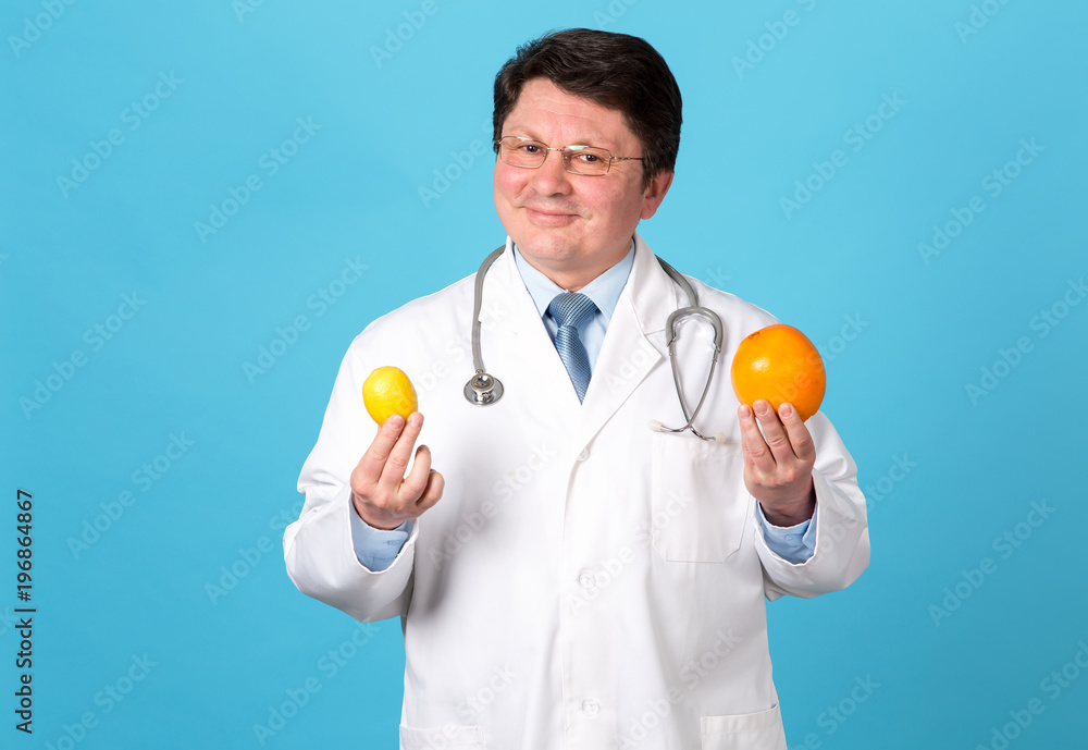 Male doctor holds fruit in hands. Vitamins for your health. Middle aged man doctor in white uniform stretching out lemon and orange fruit, smiling, while standing against blue background. Male doctor holds fruit in hands. Vitamins for your health. Middle aged man doctor in white uniform stretching out lemon and orange fruit, smiling, while standing against blue background.