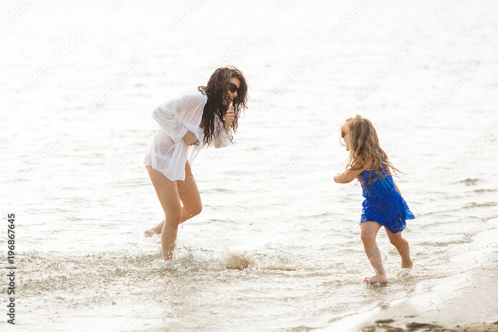 Mother and her little daughter having fun at the coast. Young pretty mom and her child playing near the water