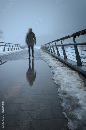 Silhouette of a man passing through a pedestrian bridge and reflected in puddles obscure in dense fog . Mystic background