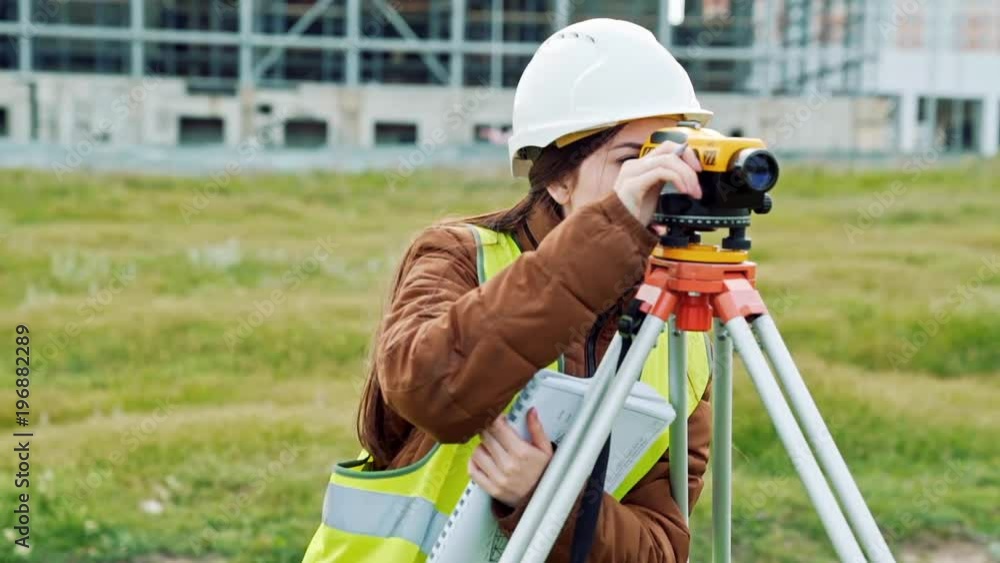 A young woman surveyor in work clothes and helmet adjusts the equipment and produces calculations on the construction site. The concept of landscape design, geodesy A young woman surveyor in work clothes and helmet adjusts the equipment and produces calculations on the construction site. The concept of landscape design, geodesy