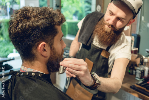 Close-up side view of a handsome redhead young man sitting on the chair of a trendy barbershop for trimming and styling his beard