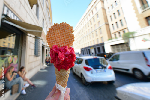 Photography Appetizing pink berry ice cream in waffle cone in female hand over Rome city view