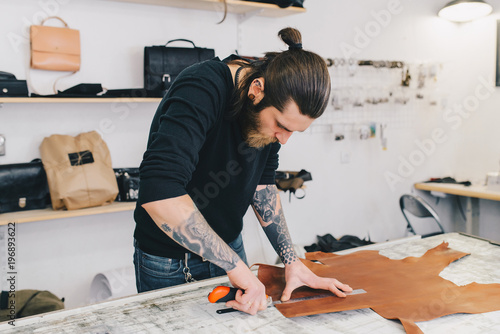 Portrait of modern handsome craftsman focused on his work, taking measurements from rough leather in studio