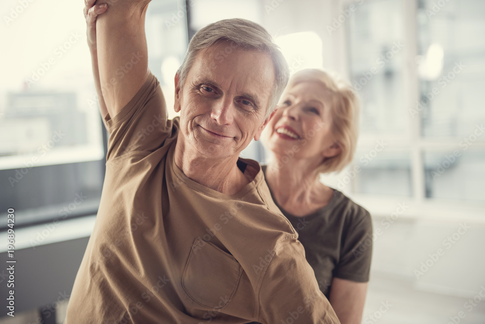Portrait of glad slim pensioner with raised arm. Smiling senior woman on background holding his arm. Focus on man Portrait of glad slim pensioner with raised arm. Smiling senior woman on background holding his arm. Focus on man