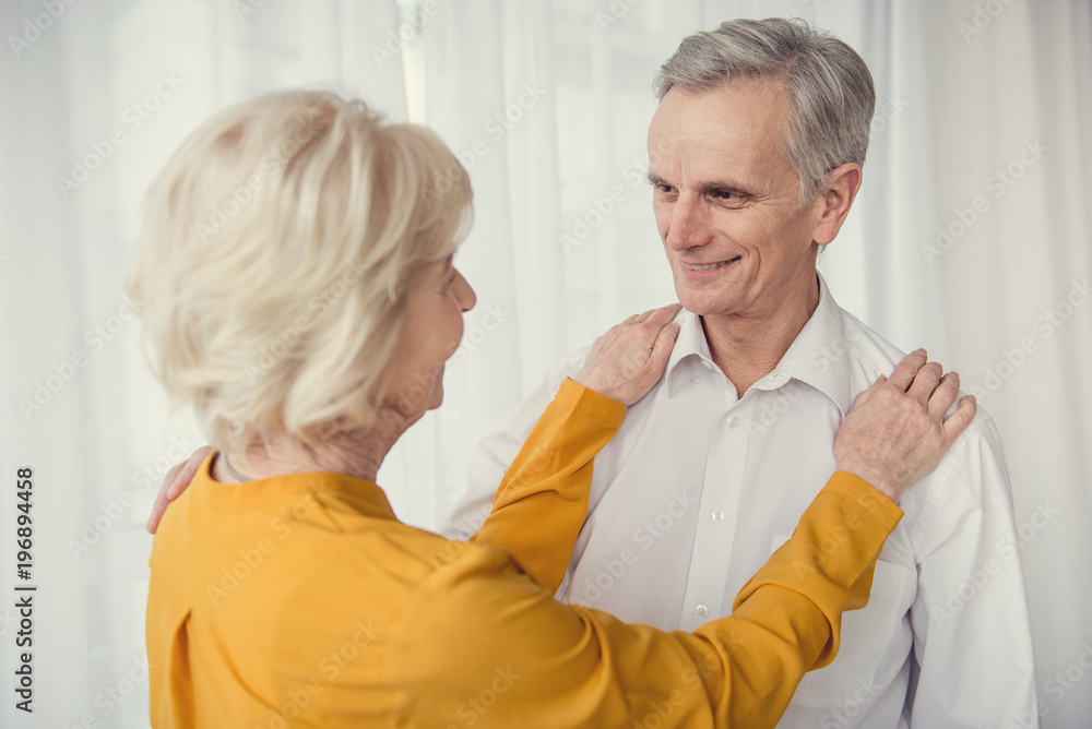Caring female pensioner looking at her smiling elderly husband, her hands lying on his shoulders. White curtain on background Caring female pensioner looking at her smiling elderly husband, her hands lying on his shoulders. White curtain on background