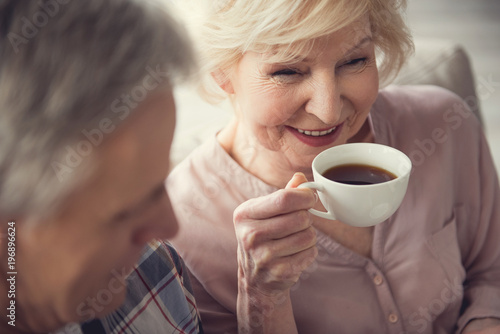 Delighted female pensioner enjoying hot drink while sitting with her husband inside. Focus on granny