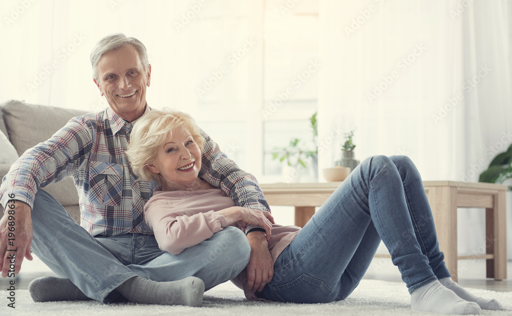 Sweet home. Portrait of cheerful senior couple resting on soft carpet in living room. They are looking at camera and laughing