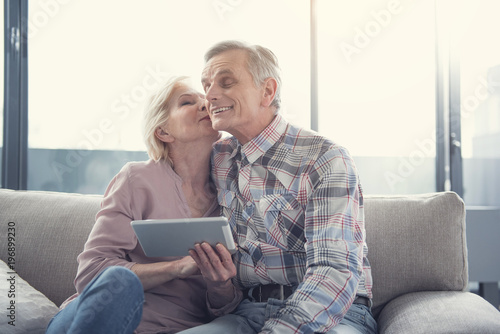 Love forever. Happy elderly woman with tablet in hand kissing her husband while sitting in living room