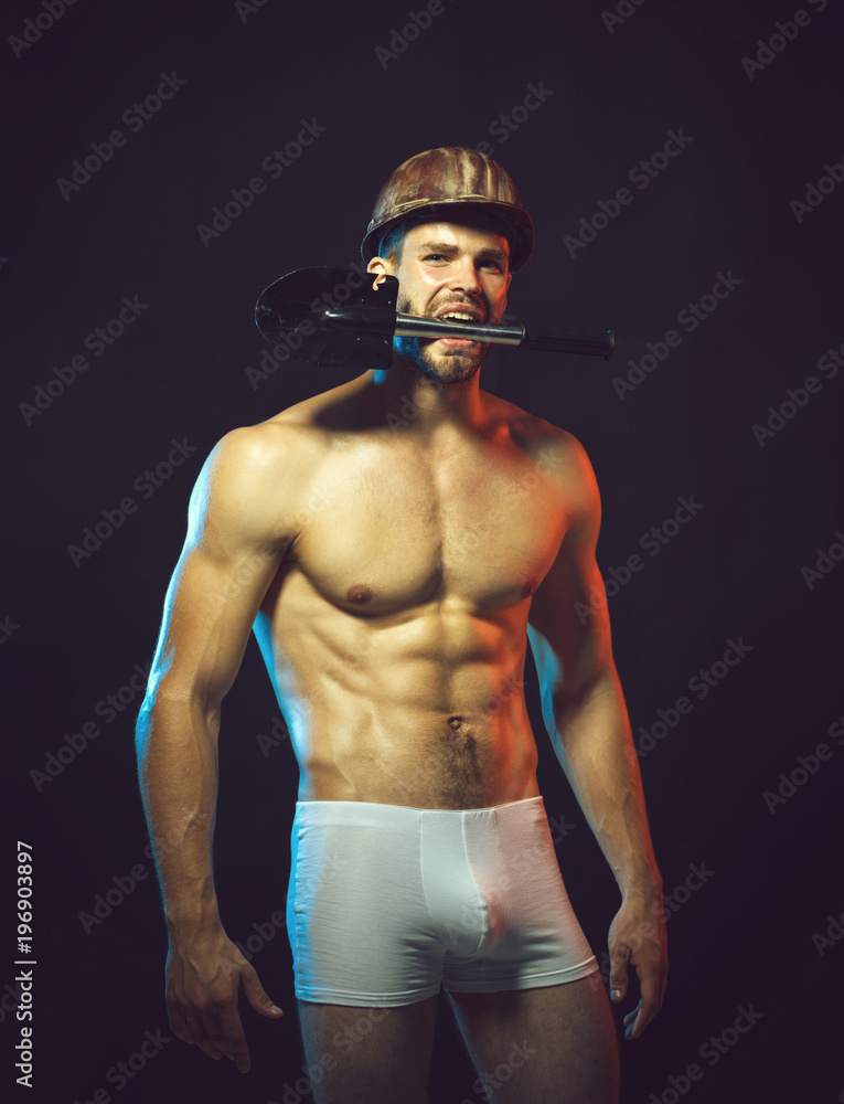 Repair, male worker in helmet holds sapper shovel in teeth, sexy builder. Construction, work and mining concept -muscular sexy professional builder, engineer, repairman in protective helmet with spade