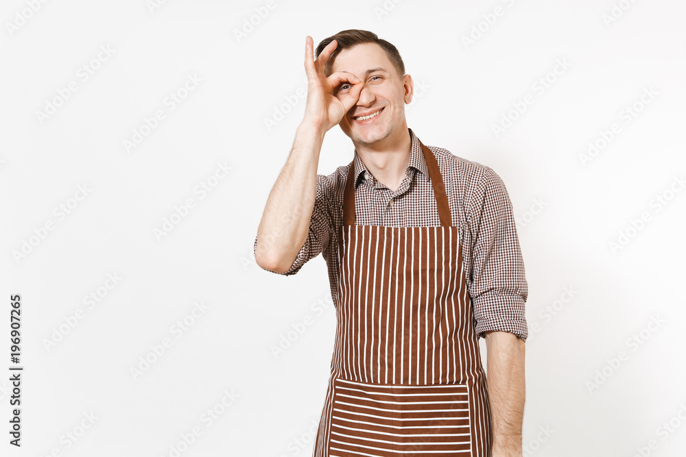 Young smiling man chef or waiter in striped brown apron, shirt showing OK gesture at eye isolated on white background. Male housekeeper or houseworker looking camera. Domestic worker for advertisement Young smiling man chef or waiter in striped brown apron, shirt showing OK gesture at eye isolated on white background. Male housekeeper or houseworker looking camera. Domestic worker for advertisement