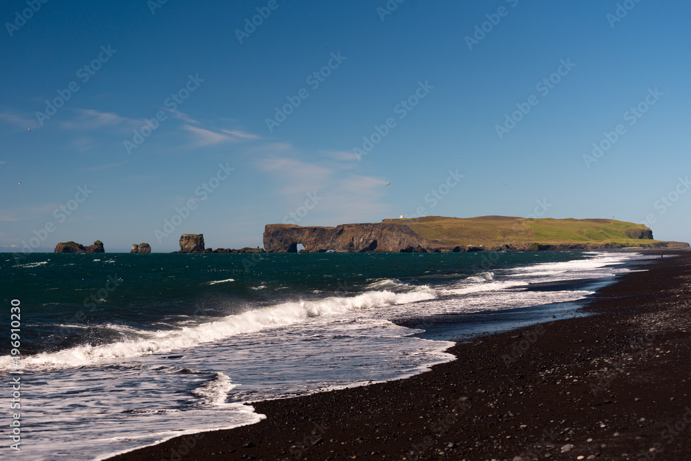 Volcanic rock on black sand beach at Vik in southern Iceland with eroded stone arch visible in distance