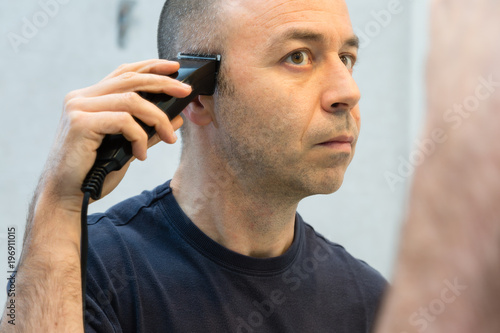 Bald Man with Black Shirt Shaving his Hair with Electric Razor in front of a Mirror