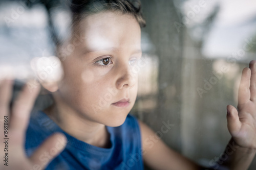 one sad little boy standing near the window at the day time.