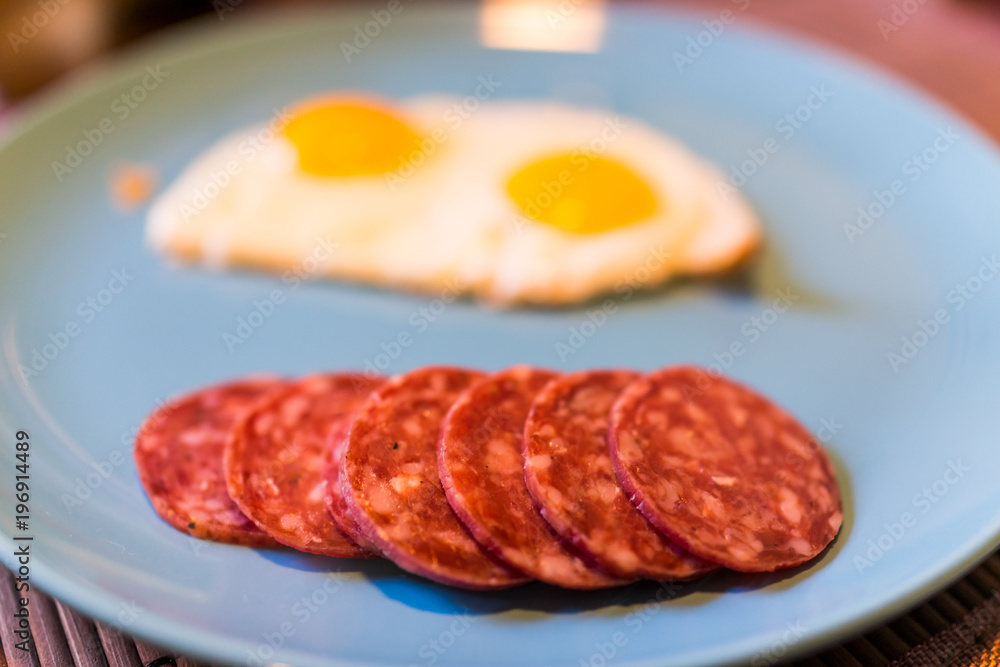 Closeup of two fried eggs on large blue plate with cured meat hard salami slices Closeup of two fried eggs on large blue plate with cured meat hard salami slices