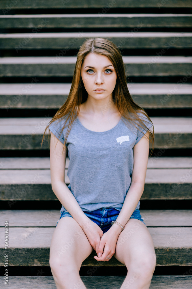 Fototapeta premium Young girl portrait. Blue eyed brunette female expressive sensual face. Big sad innocent eyes of adorable woman sitting on wooden stairs outdoor. Reflexive person state of mind. Deep inner mood.