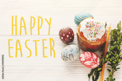 happy easter text. season's greetings card. stylish painted eggs and easter cake on white rustic wooden background with spring flowers and candle. modern easter image