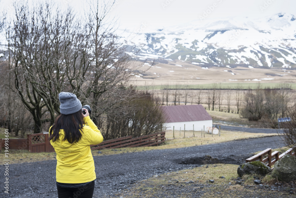 Long hair young asian lady in yellow warm shirt use camera capture photo of Icelandic snowcapped  mountain view in winter.