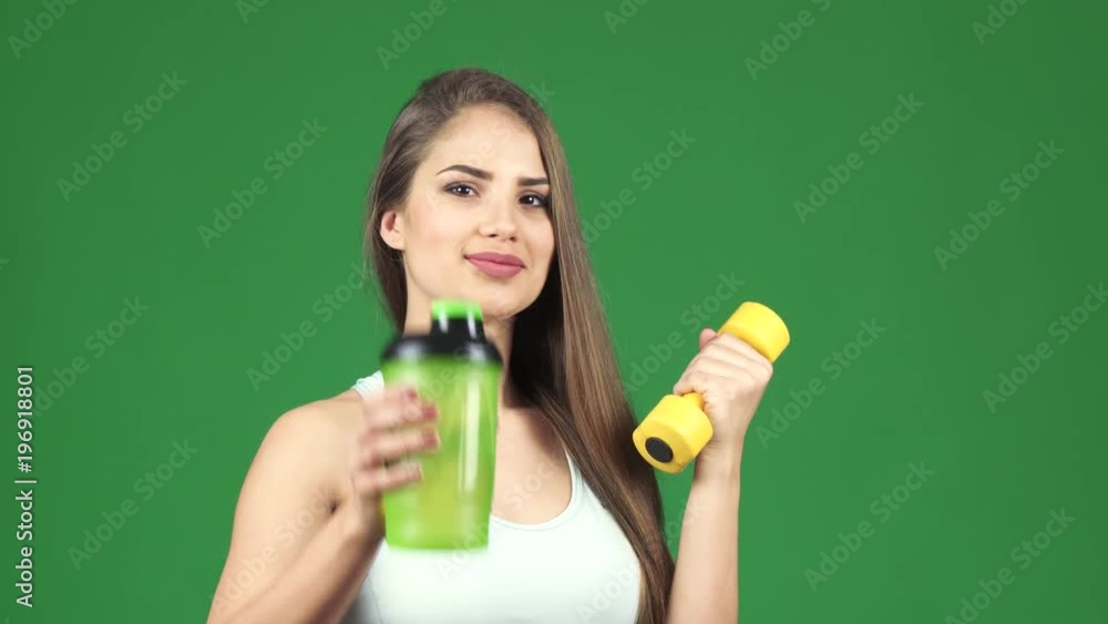 Cheerful young beautiful athletic woman smiling joyfully holding out a bottle of water to the camera posing with a dumbbell on green chromakey background sports dieting healthy lifestyle. Cheerful young beautiful athletic woman smiling joyfully holding out a bottle of water to the camera posing with a dumbbell on green chromakey background sports dieting healthy lifestyle.