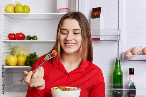 Happy lovely woman eats fresh vegetrian salad made with cucumbers and tomatoes, stands near opened fridge full of vegetables and fruits, being vegan, doesn`t eat meat. Pretty female with dish photo