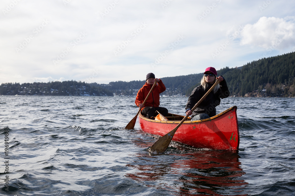 Obraz premium Couple friends on a wooden canoe are paddling in an inlet surrounded by Canadian mountains. Taken in Indian Arm, near Deep Cove, North Vancouver, British Columbia, Canada.