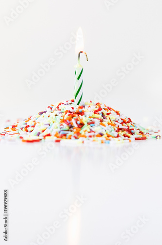 Burning white and green striped spiral birthday candle for dopping cakes, cupcakes or muffins as decoration on pile of colorful sugar spinkles on white background photo