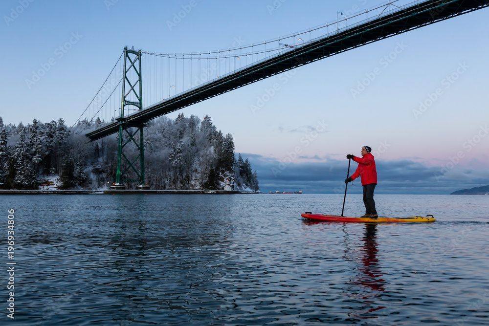 Naklejka premium Adventurous man on a Standup Paddle Board is padding near Lions Gate Bridge during a vibrant winter sunrise. Taken in Vancouver, British Columbia, Canada.