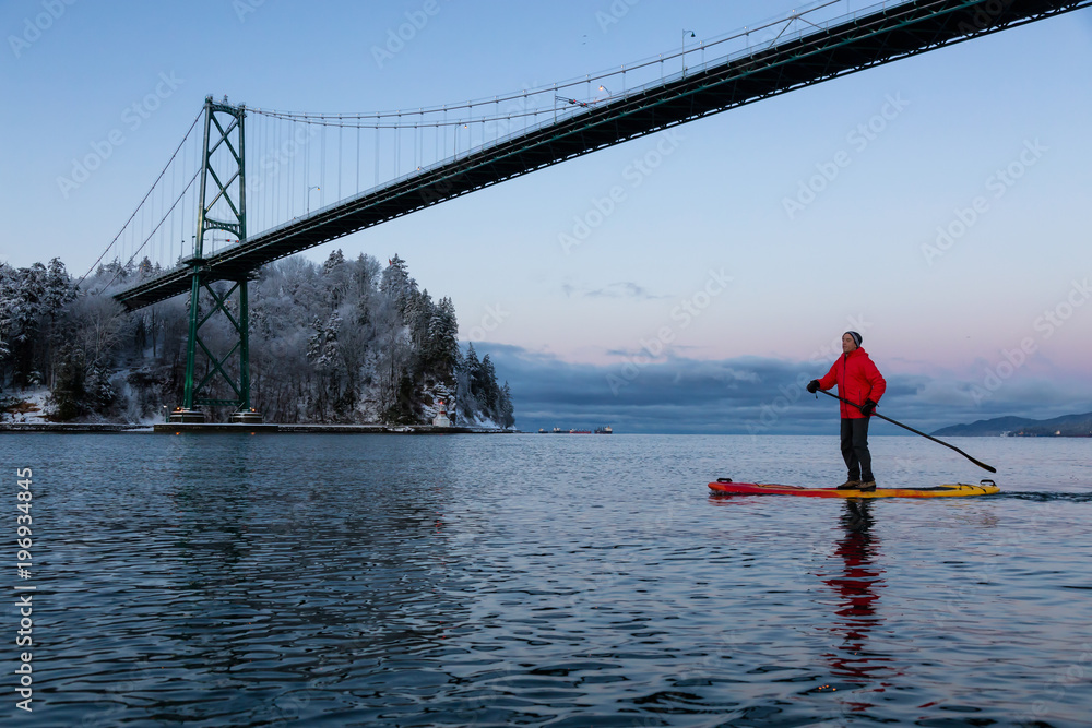 Adventurous man on a Standup Paddle Board is padding near Lions Gate Bridge during a vibrant winter sunrise. Taken in Vancouver, British Columbia, Canada.